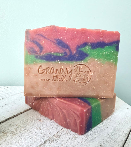 Two handmade soap bars stacked on a white wooden surface against a pale blue wall. The top bar is labeled 'Granny Hen Soap Company' and features a marbled pattern of pink, purple, green, and beige.
