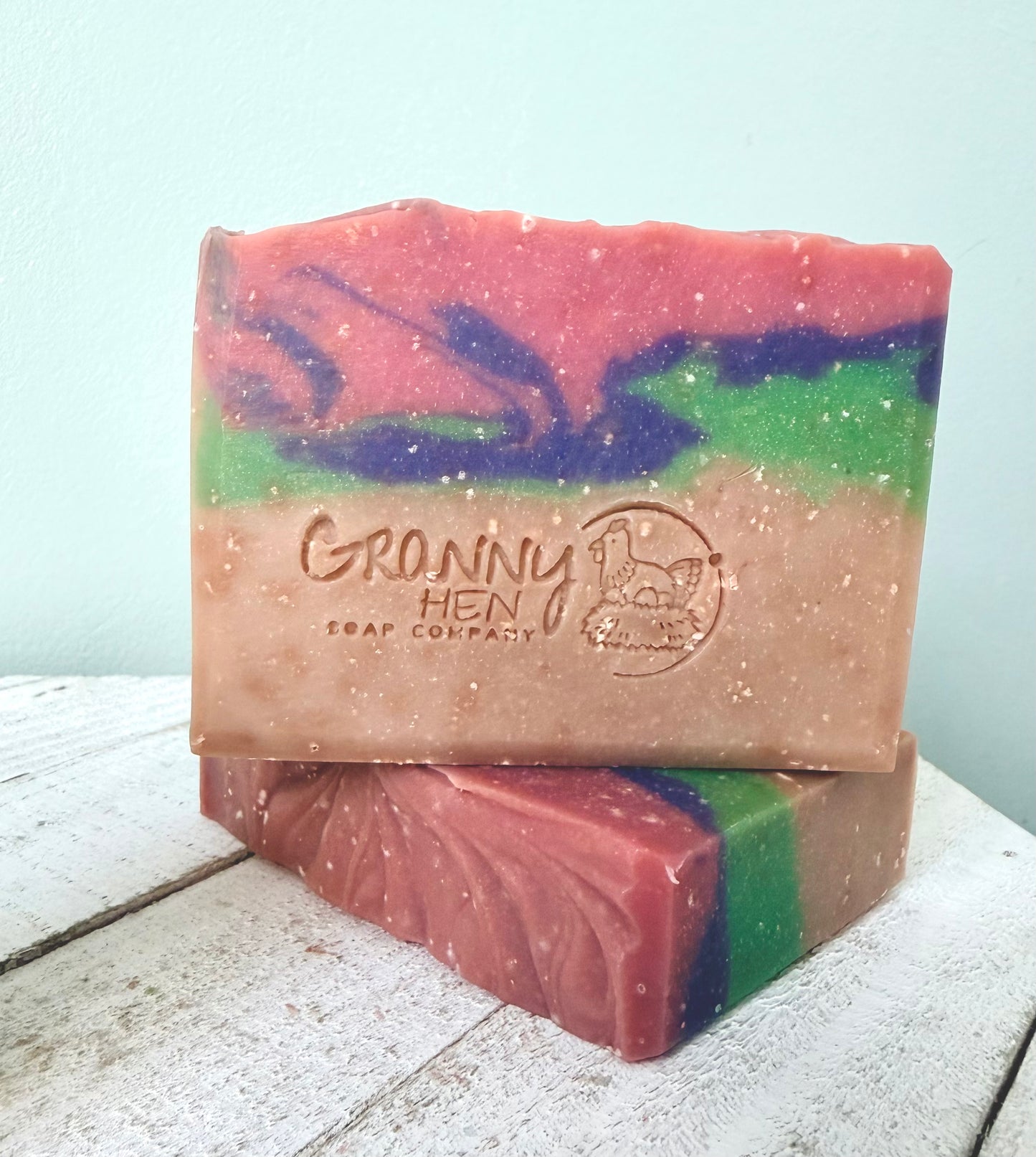 Two handmade soap bars stacked on a white wooden surface against a pale blue wall. The top bar is labeled 'Granny Hen Soap Company' and features a marbled pattern of pink, purple, green, and beige.