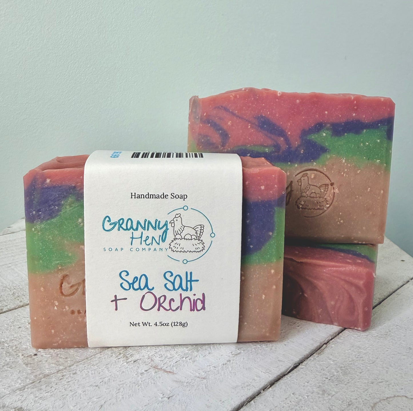 Three handmade soap bars with a white paper label reading 'Granny Hen Soap Company Sea Salt + Orchid' on a rustic white wooden surface against a pale blue wall.