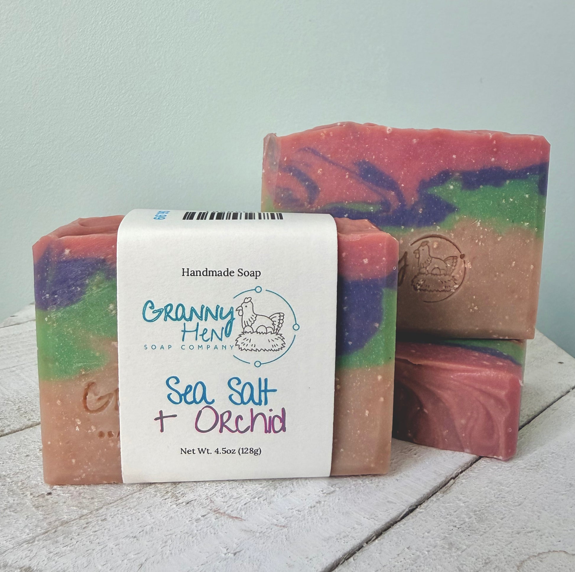 Three handmade soap bars with a white paper label reading 'Granny Hen Soap Company Sea Salt + Orchid' on a rustic white wooden surface against a pale blue wall.