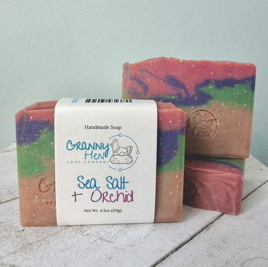 Three handmade soap bars with a white paper label reading 'Granny Hen Soap Company Sea Salt + Orchid' on a rustic white wooden surface against a pale blue wall.